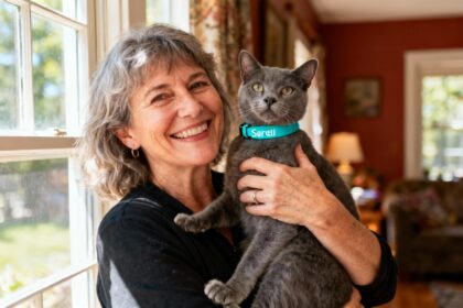 Femme de 55 ans souriante tenant un chat gris portant un collier Seresto turquoise dans un intérieur chaleureux